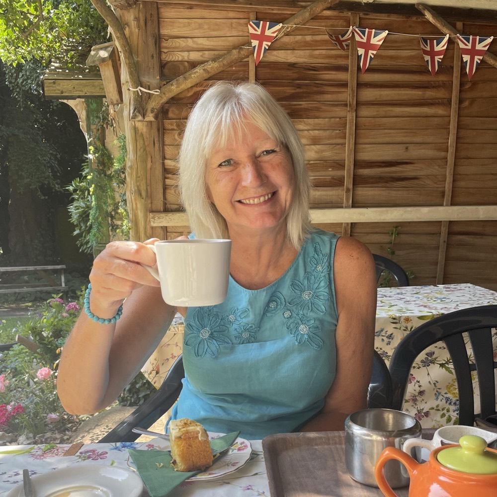 Dawn Mitchell smiling in a sunny garden, holding a white mug, in a turquoise dress, with red-white-and-blue bunting strung along a wooden cabin behind her.