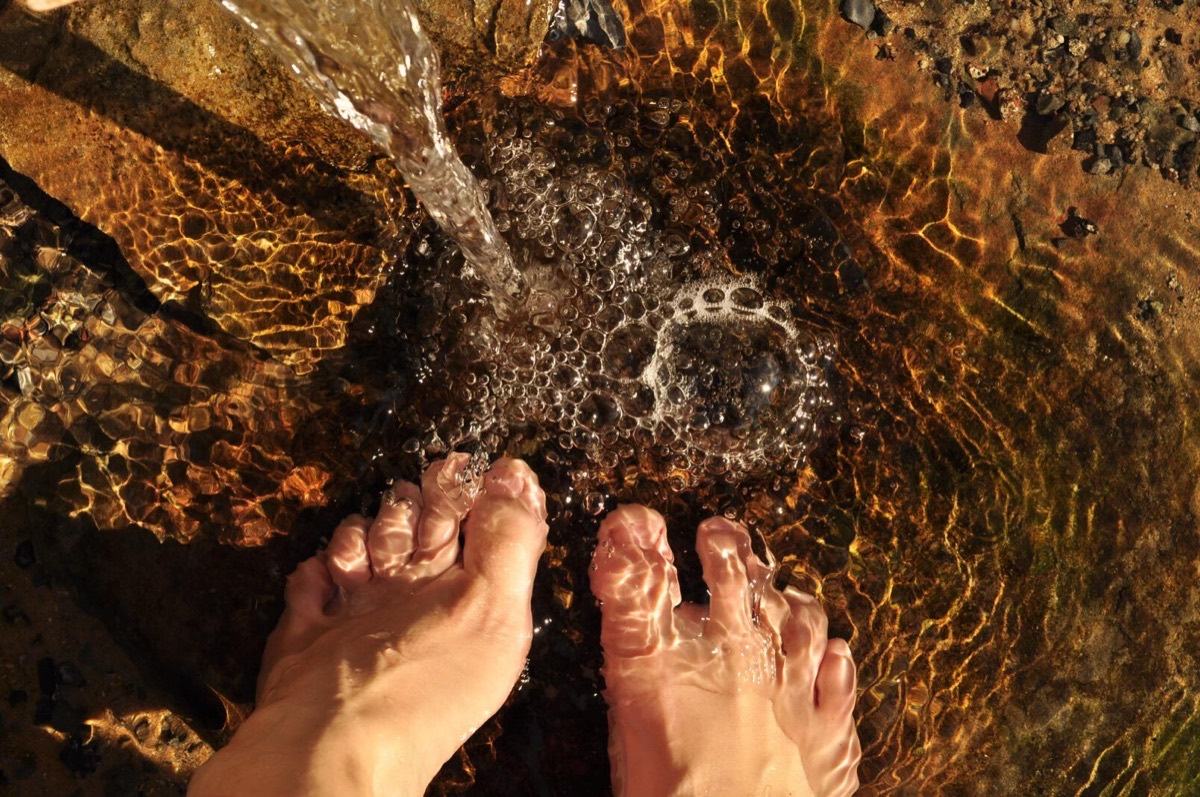 Two bare feet standing in a shallow, sunlit stream, seen from above, with clear water pouring over pebbles and rock.